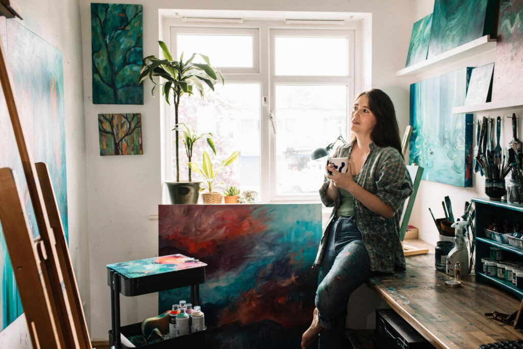 Artist sitting on desk in studio holding a mug. Colourful abstract art, paintbrushes, an easel and plants are visible.