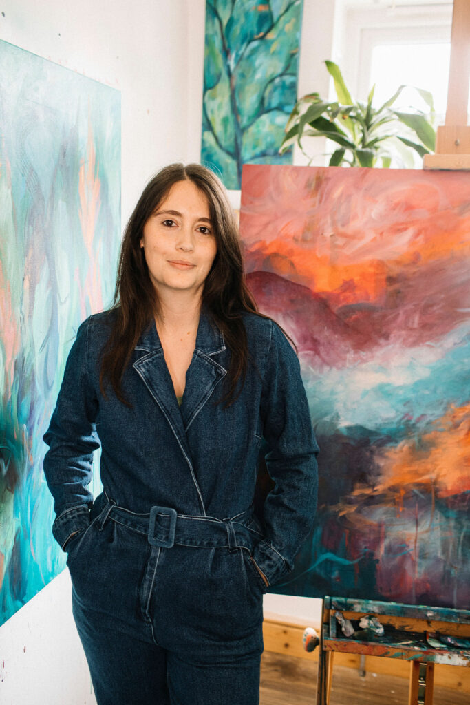 Artist in denim jumpsuit standing next to colourful abstract landscape painting, a small tree painting and plant are in the background.