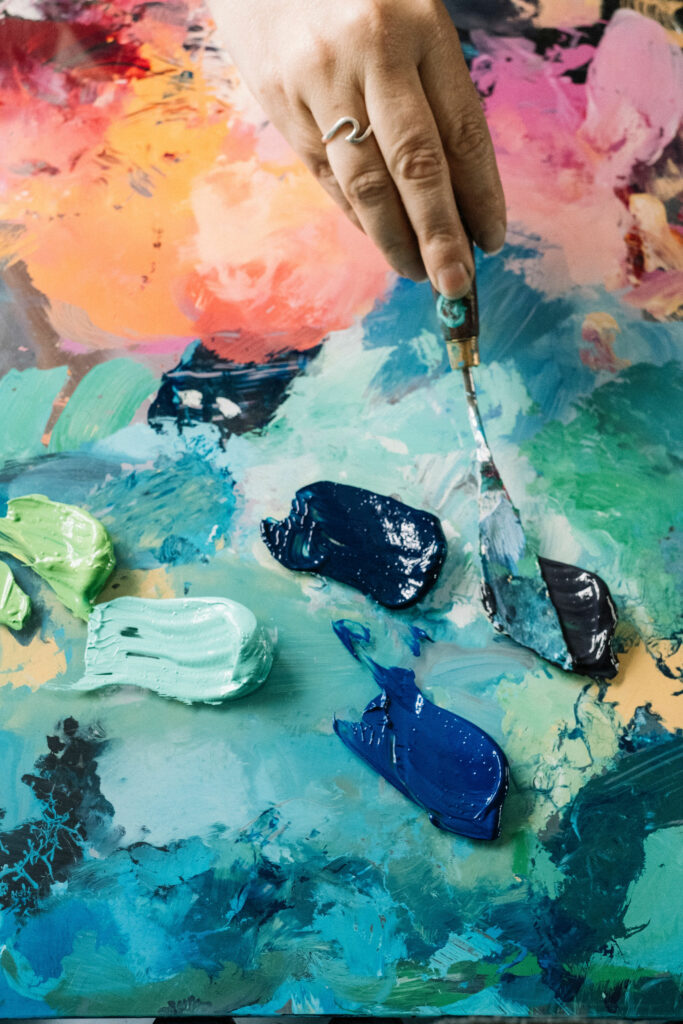 Artist's hand holding a palette knife putting thick paint onto a glass plaette with orange and blues